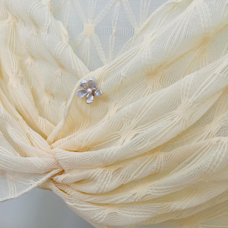 May include: Close-up of a cream-colored, textured shawl with a floral brooch. The shawl is draped and knotted, showcasing a delicate, woven pattern. The brooch is silver-toned with a pearl-like center, adding a touch of elegance.
