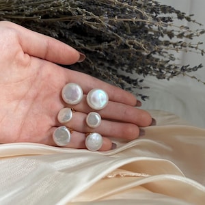 May include: Five white pearl earrings with gold settings. The earrings are arranged on a hand with a light pink background.