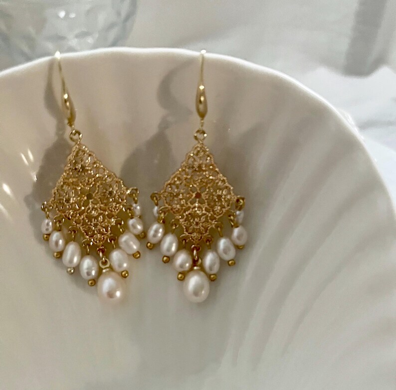 Peut inclure: Une paire de boucles d'oreilles pendantes dor&eacute;es avec un &eacute;l&eacute;ment en m&eacute;tal filigran&eacute; en forme de losange. Chaque boucle est orn&eacute;e de plusieurs petites perles d'eau douce blanches de forme ovale suspendues &agrave; la base.