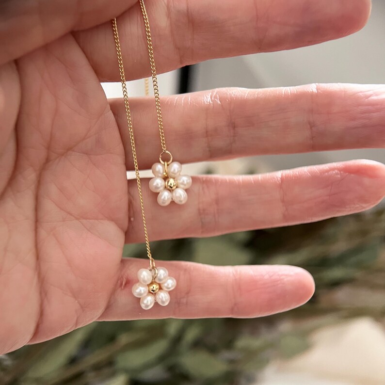 May include: A delicate gold chain necklace with two pearl flower charms. Each flower charm is made of small white pearls arranged in a circular pattern.