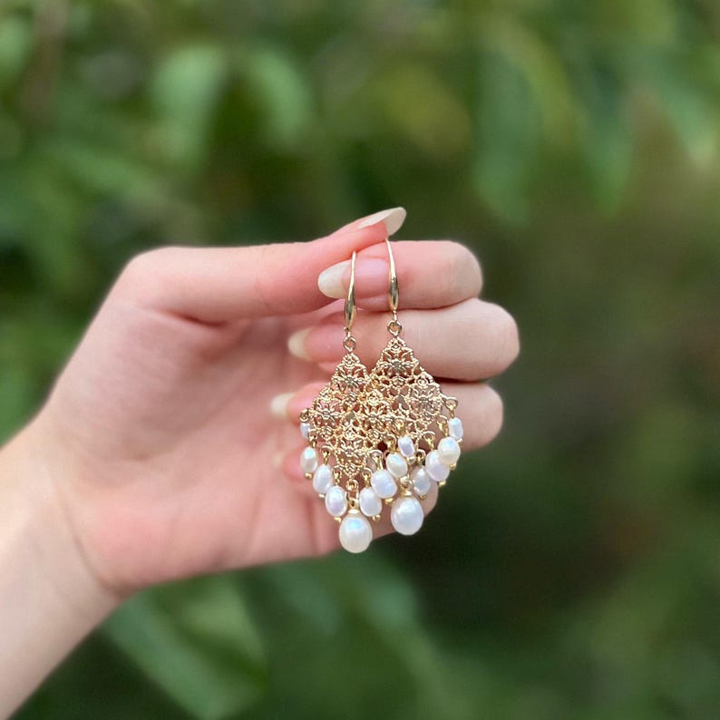 Peut inclure: Une paire de boucles d'oreilles chandelier dor&eacute;es avec un design en m&eacute;tal filigran&eacute; complexe. Les boucles d'oreilles sont orn&eacute;es de plusieurs petites perles blanches de forme ovale qui pendent au bas de la monture m&eacute;tallique triangulaire.