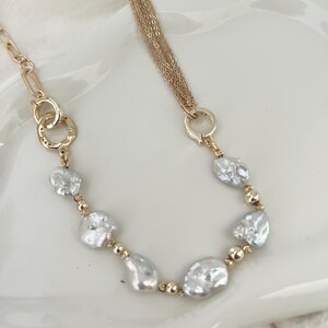 Peut inclure: Un collier dor&eacute; orn&eacute; de perles blanches irr&eacute;guli&egrave;res et de petites perles dor&eacute;es. Le collier comporte une cha&icirc;ne &agrave; plusieurs brins d'un c&ocirc;t&eacute; et une cha&icirc;ne avec des cercles imbriqu&eacute;s de l'autre.