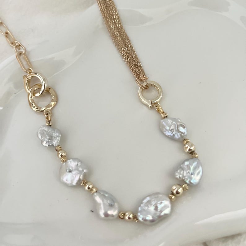 Peut inclure: Un collier dor&eacute; orn&eacute; de perles blanches irr&eacute;guli&egrave;res et de petites perles dor&eacute;es. Le collier comporte une cha&icirc;ne &agrave; plusieurs brins d'un c&ocirc;t&eacute; et une cha&icirc;ne avec des cercles imbriqu&eacute;s de l'autre.