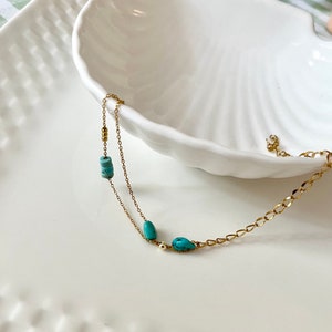 May include: A delicate gold chain necklace with two turquoise stones and small gold beads. The necklace is displayed on a white seashell-shaped dish.
