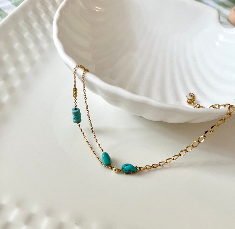 May include: A delicate gold chain necklace with two turquoise stones and small gold beads. The necklace is displayed on a white seashell-shaped dish.