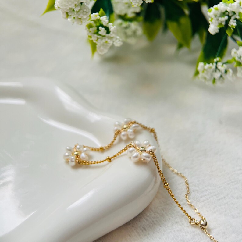 May include: A delicate gold chain necklace with small pearl flower accents. The necklace is on a white surface.