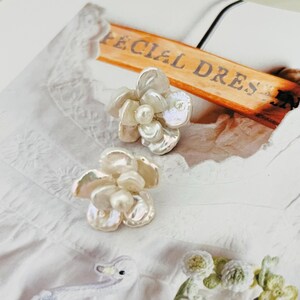 Op de afbeelding: Twee bloemvormige oorbellen met iriserende bloemblaadjes en een centrale parel. De oorbellen zijn geplaatst tegen een witte achtergrond met de tekst "Fecial Dres" zichtbaar op een houten bord.