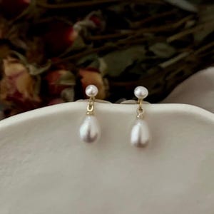 Peut inclure: Une paire de boucles d'oreilles pendantes en perles dorées. Chaque boucle d'oreille présente une petite perle en haut et une perle plus grosse en forme de larme qui pend en dessous.