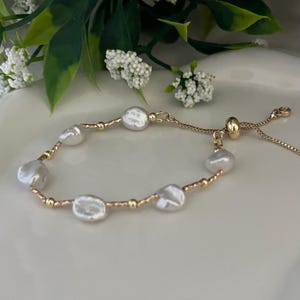 Pode incluir: Pulseira dourada com seis pérolas brancas de formato irregular. Pequenas contas douradas e uma delicada corrente dourada conectam as pérolas. A pulseira tem um fecho ajustável.