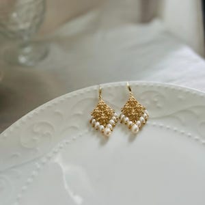 Peut inclure: Une paire de boucles d'oreilles pendantes dor&eacute;es avec une pi&egrave;ce centrale en m&eacute;tal textur&eacute; en forme de losange, orn&eacute;e de petites perles blanches ovales suspendues au bord inf&eacute;rieur par de fins crochets dor&eacute;s.