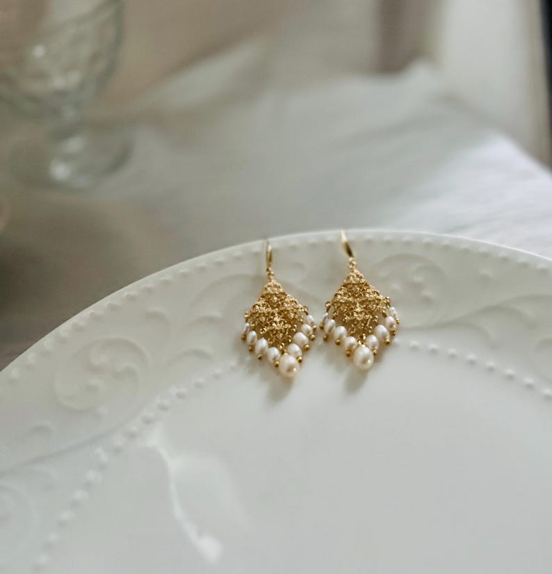 Peut inclure: Une paire de boucles d'oreilles pendantes dor&eacute;es avec une pi&egrave;ce centrale en m&eacute;tal textur&eacute; en forme de losange, orn&eacute;e de petites perles blanches ovales suspendues au bord inf&eacute;rieur par de fins crochets dor&eacute;s.