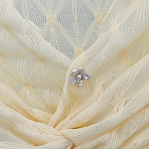 May include: Close-up of a pale yellow knit top with a delicate floral brooch. The fabric has a subtle diamond pattern. The brooch is silver-toned with a cluster of petal-shaped elements.