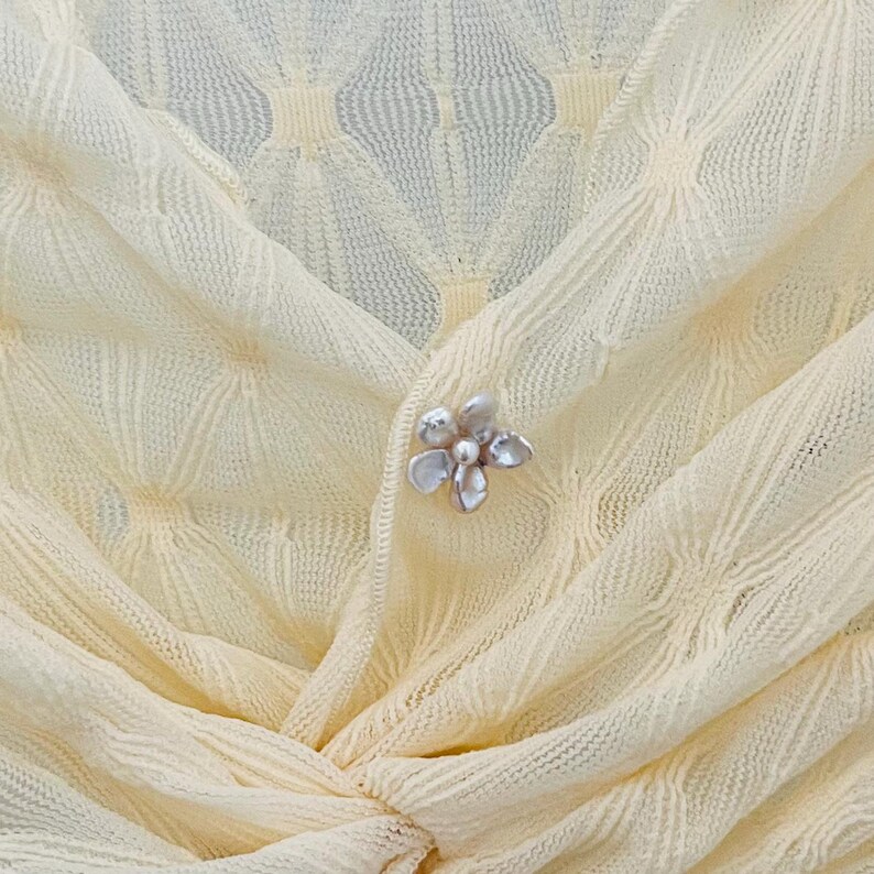 May include: Close-up of a pale yellow knit top with a delicate floral brooch. The fabric has a subtle diamond pattern. The brooch is silver-toned with a cluster of petal-shaped elements.