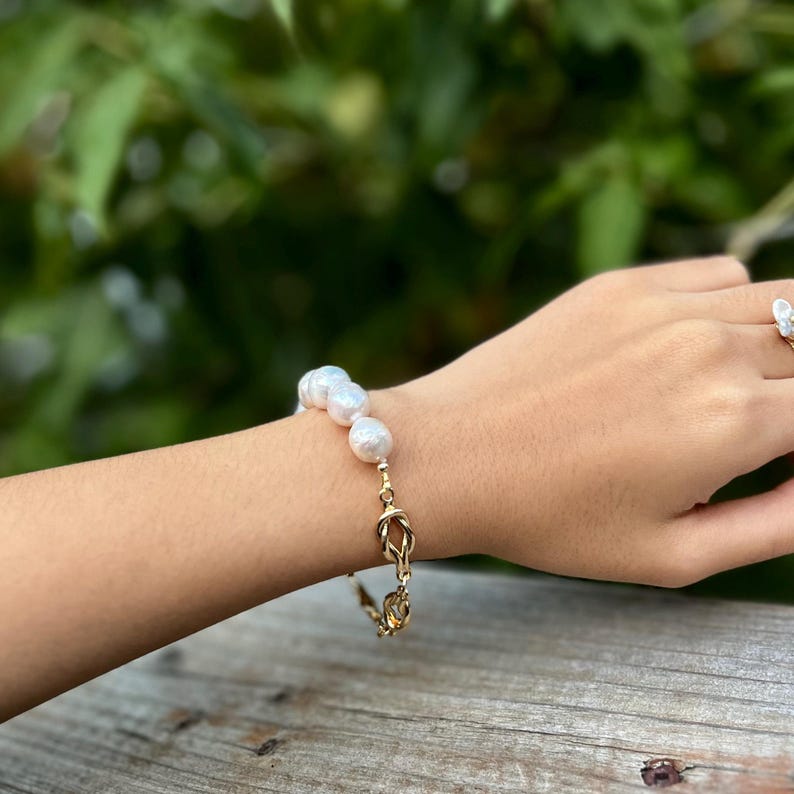 Puede incluir: Un brazalete de cadena dorada con un dise&ntilde;o de nudo y una fila de perlas blancas.