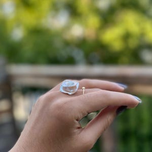 May include: A silver ring with a large, irregular, white pearl-like stone. The ring is on a person's finger.