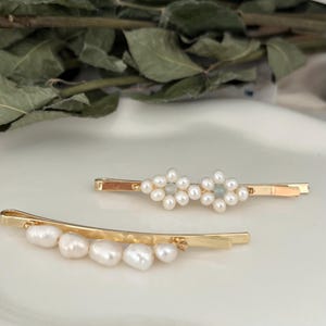 Puede incluir: Dos horquillas doradas. Una presenta un dise&ntilde;o floral de perlas blancas, la otra una fila de perlas blancas. Los accesorios para el cabello se colocan sobre un fondo suave y neutro.
