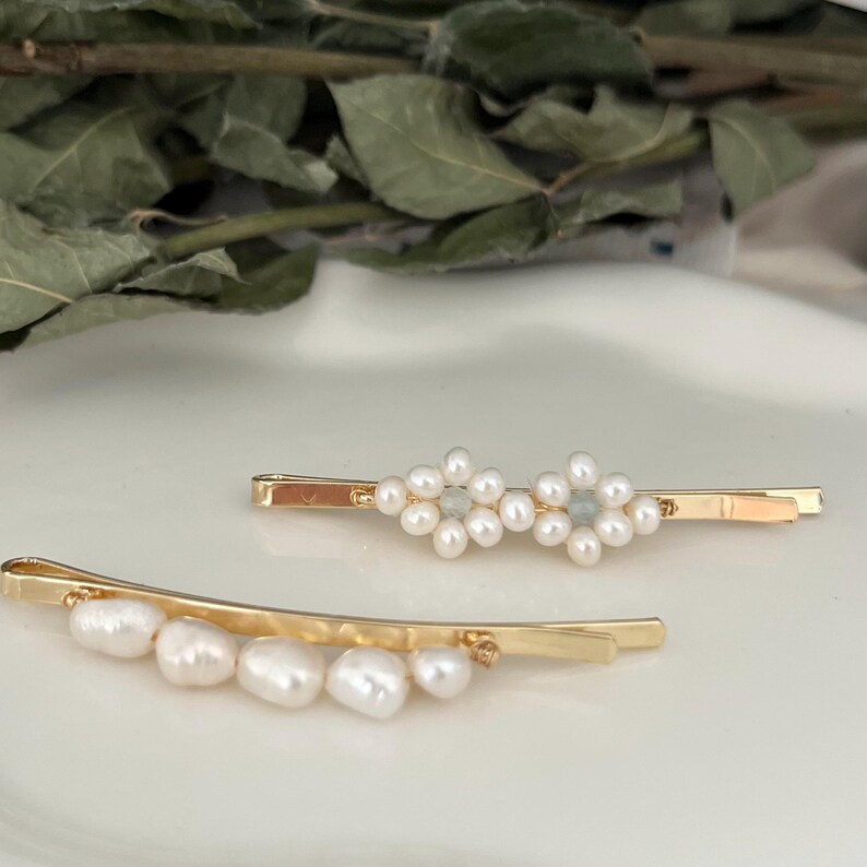 Puede incluir: Dos horquillas doradas. Una presenta un dise&ntilde;o floral de perlas blancas, la otra una fila de perlas blancas. Los accesorios para el cabello se colocan sobre un fondo suave y neutro.