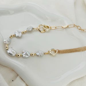Peut inclure: Un collier dor&eacute; avec des perles blanches irr&eacute;guli&egrave;res et de petites perles dor&eacute;es. Le collier est dot&eacute; d'un fermoir &agrave; double boucle et d'une cha&icirc;ne d&eacute;licate avec une frange de brins dor&eacute;s. Le bijou est pr&eacute;sent&eacute; sur une surface blanche.
