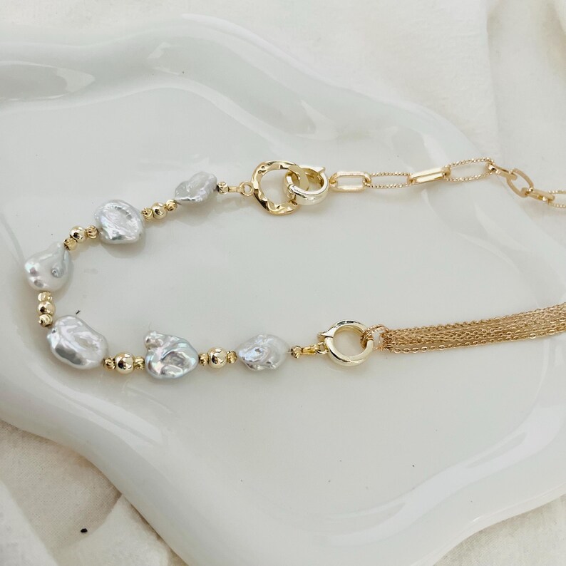 Peut inclure: Un collier dor&eacute; avec des perles blanches irr&eacute;guli&egrave;res et de petites perles dor&eacute;es. Le collier est dot&eacute; d'un fermoir &agrave; double boucle et d'une cha&icirc;ne d&eacute;licate avec une frange de brins dor&eacute;s. Le bijou est pr&eacute;sent&eacute; sur une surface blanche.