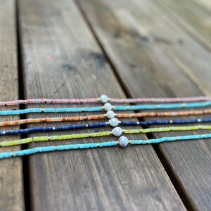 May include: A set of six delicate necklaces featuring a variety of colorful beads and a single pearl accent. The necklaces are arranged in a rainbow pattern on a wooden surface.
