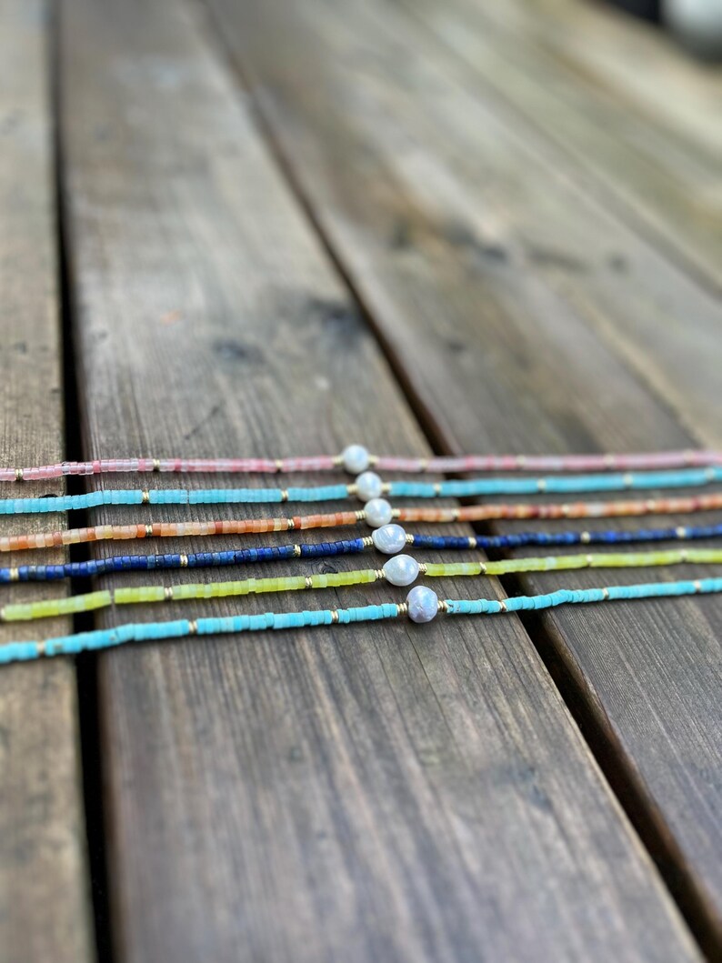 May include: A set of six delicate necklaces featuring a variety of colorful beads and a single pearl accent. The necklaces are arranged in a rainbow pattern on a wooden surface.