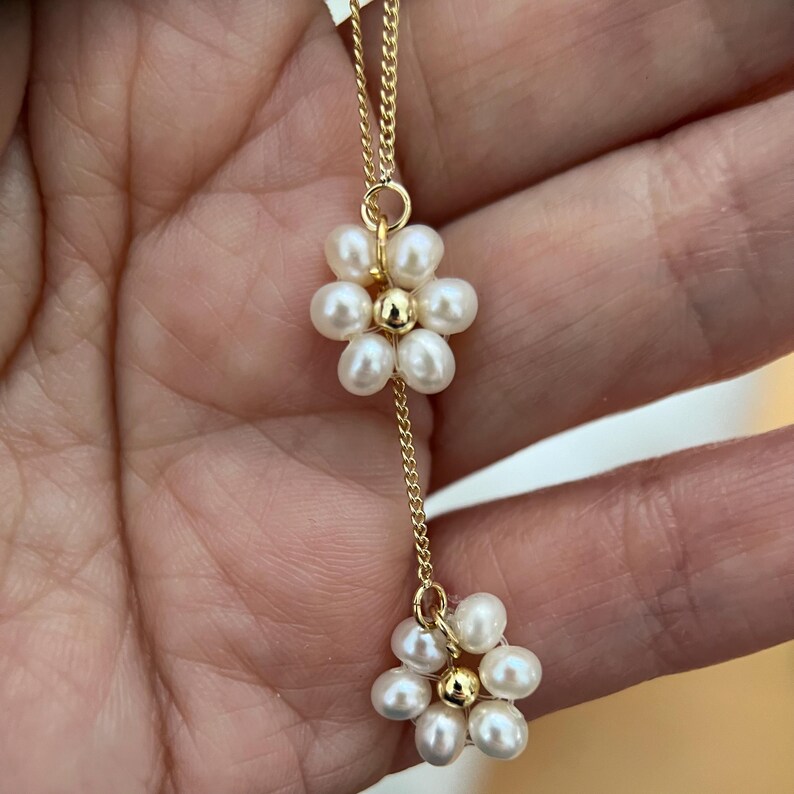 May include: A delicate gold chain necklace with two pearl flower charms. Each flower charm is made of small white pearls arranged in a circular pattern.