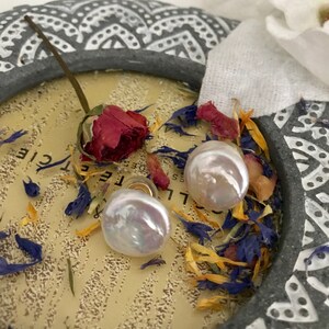 May include: Two white pearl earrings with a gold backing, displayed on a decorative surface with dried flowers.