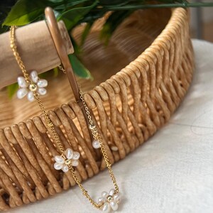 May include: A delicate gold chain necklace with three small pearl flower charms. The necklace is displayed on a woven basket.