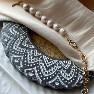 Puede incluir: Un brazalete de cadena dorada con un dise&ntilde;o anudado y una fila de perlas blancas.