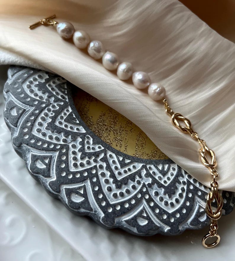 Puede incluir: Un brazalete de cadena dorada con un dise&ntilde;o anudado y una fila de perlas blancas.