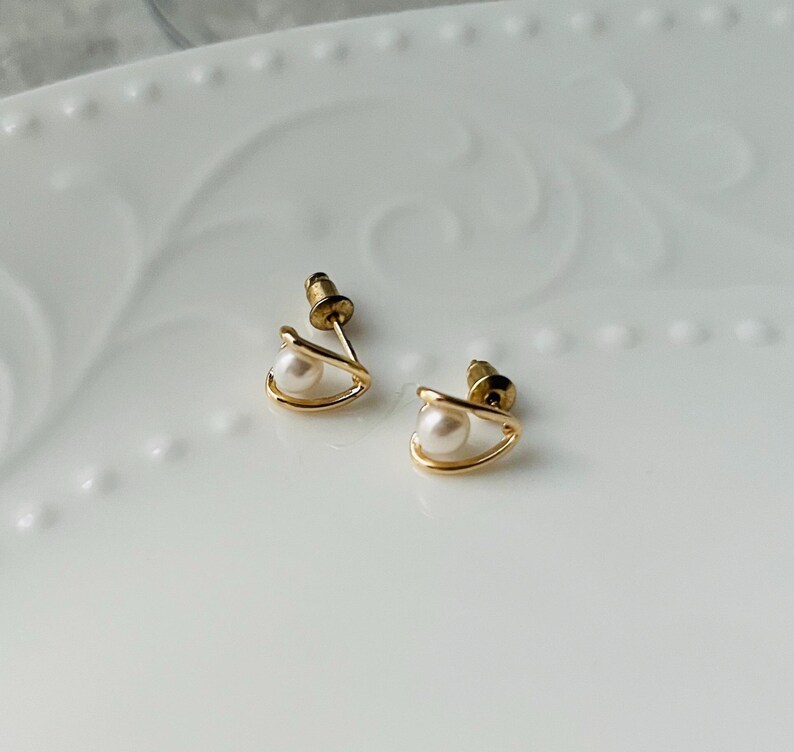K&ouml;nnte beinhalten: Ein Paar goldfarbene Ohrringe mit je einer Perle. Die Ohrringe haben ein schlichtes, modernes Design.