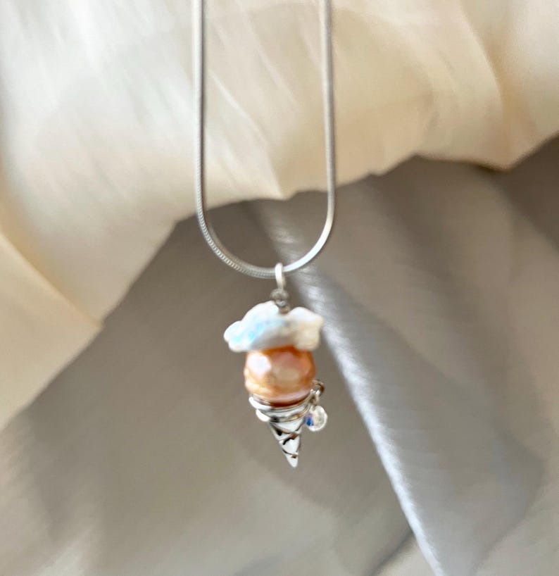 Peut inclure: Un collier en cha&icirc;ne argent avec un pendentif en forme de cornet de glace en argent et en perle. La glace est de couleur rose p&acirc;le avec un tourbillon blanc sur le dessus.