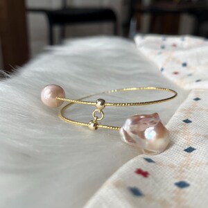 Peut inclure: Un bracelet jonc en or avec deux brins d&eacute;licats et un charme en perle &agrave; chaque extr&eacute;mit&eacute;. Les perles sont de couleur rose p&acirc;le et ont une forme irr&eacute;guli&egrave;re.