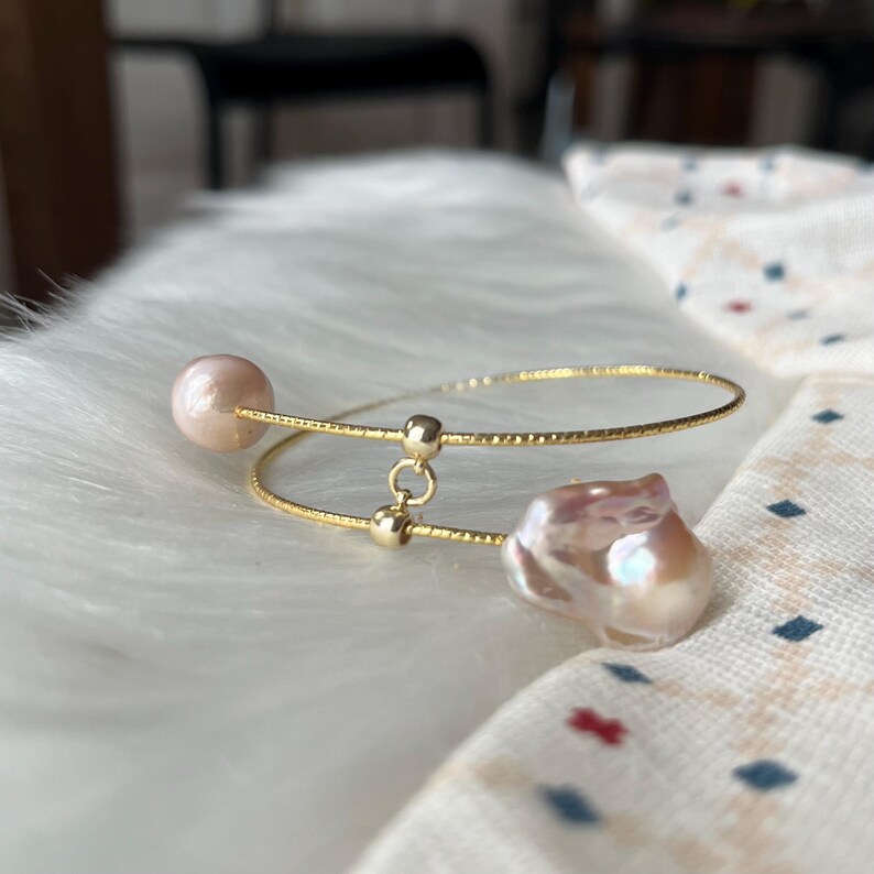 Peut inclure: Un bracelet jonc en or avec deux brins d&eacute;licats et un charme en perle &agrave; chaque extr&eacute;mit&eacute;. Les perles sont de couleur rose p&acirc;le et ont une forme irr&eacute;guli&egrave;re.