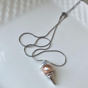 Peut inclure: Un collier en cha&icirc;ne argent&eacute;e avec un pendentif en forme de cornet de glace. Le cornet est rempli de deux perles, une blanche et une rose.