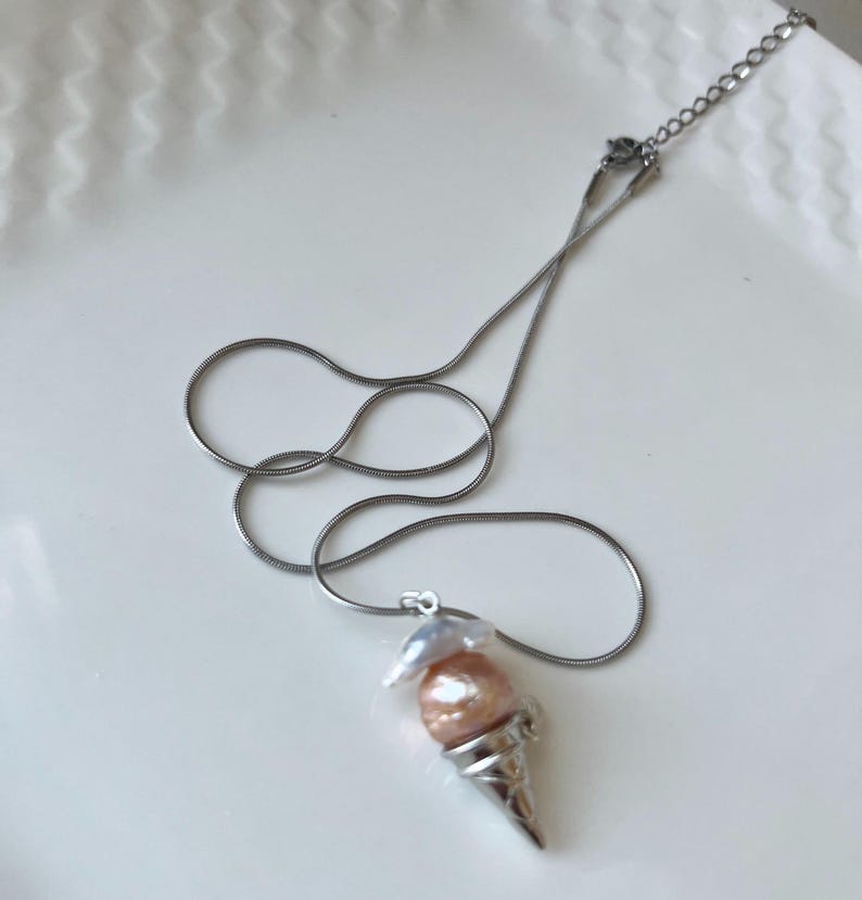 Peut inclure: Un collier en cha&icirc;ne argent&eacute;e avec un pendentif en forme de cornet de glace. Le cornet est rempli de deux perles, une blanche et une rose.