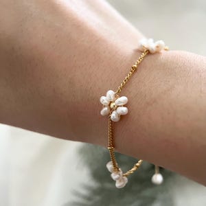 May include: A delicate gold chain bracelet featuring small white pearl flowers. The bracelet is crafted from a thin gold chain with small white pearls arranged in the shape of flowers. This bracelet is a simple and elegant piece of jewellery that would be perfect for any occasion.