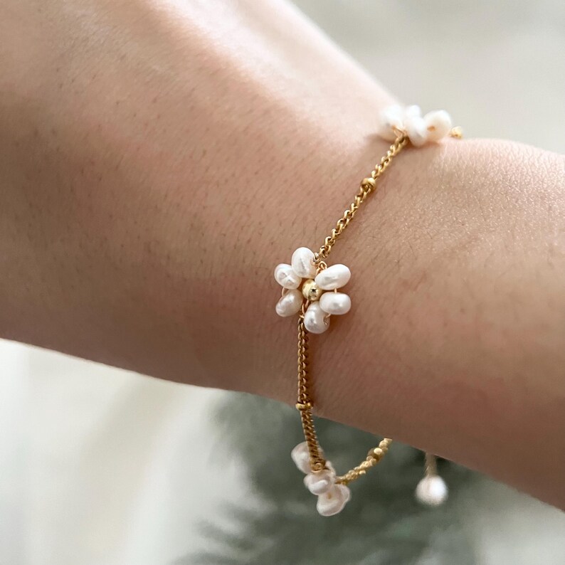 May include: A delicate gold chain bracelet featuring small white pearl flowers. The bracelet is crafted from a thin gold chain with small white pearls arranged in the shape of flowers. This bracelet is a simple and elegant piece of jewellery that would be perfect for any occasion.