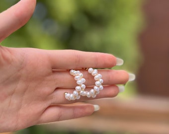 Puede incluir: Un par de pendientes de aro dorados con detalles de perlas blancas. Los pendientes están hechos de pequeñas perlas redondas que están enhebradas juntas en un bucle continuo.