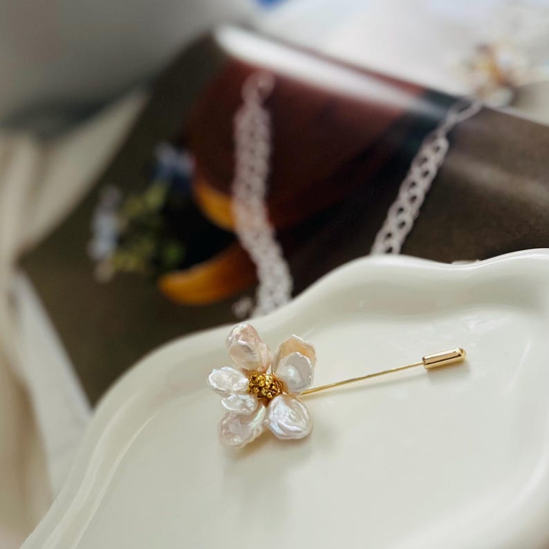 Puede incluir: Un delicado broche de flor de perla blanca con un alfiler de oro. La flor tiene un centro hecho de peque&ntilde;as cuentas doradas.