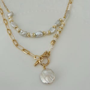May include: A two-strand necklace with a gold chain and white baroque pearls. The necklace has a gold toggle clasp and a large, irregular pearl pendant. The design is elegant and suitable for special occasions.
