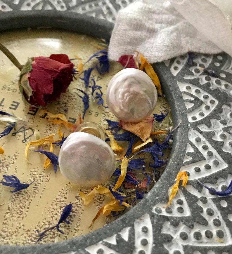 May include: A pair of pearl earrings with a white and pink iridescent finish. The earrings are displayed on a gray background with dried flowers.