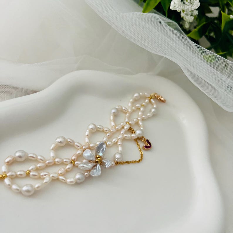 May include: A delicate pearl necklace with multiple strands and a floral accent. The necklace features small, irregularly shaped pearls in shades of white and cream, connected by gold-toned metal. A small red heart charm hangs from the necklace.