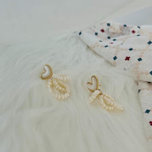 May include: A pair of gold heart-shaped earrings with white pearl drop accents. The earrings are on a white fluffy surface.