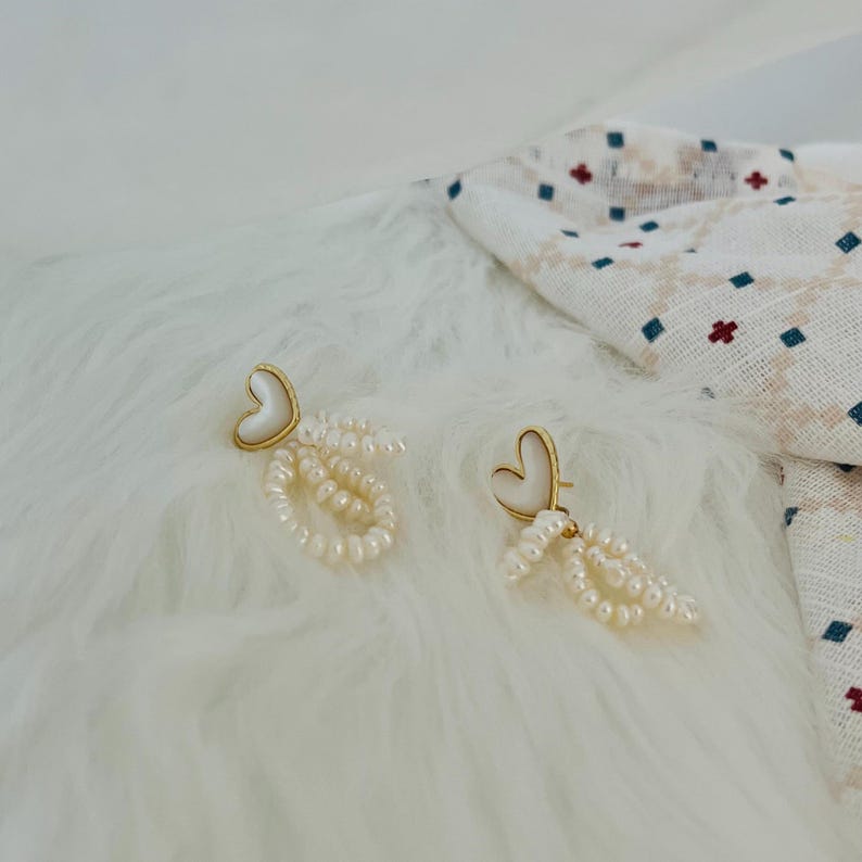 May include: A pair of gold heart-shaped earrings with white pearl drop accents. The earrings are on a white fluffy surface.