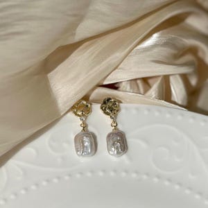 Puede incluir: Un par de pendientes de oro y perla. Los pendientes tienen un peque&ntilde;o disco de oro con una superficie texturizada y una gran perla irregular.