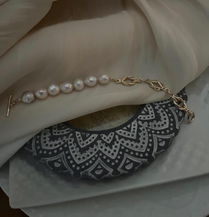 Puede incluir: Un brazalete de cadena dorada con cierre de palanca y una fila de perlas grandes y blancas.