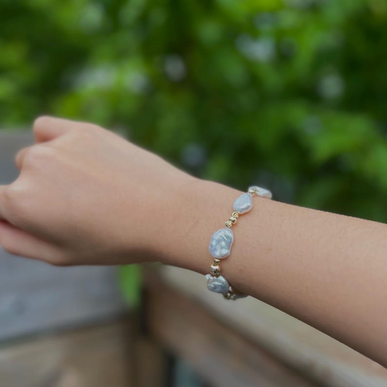 Peut inclure: Un bracelet orn&eacute; de grosses perles bleues claires irr&eacute;guli&egrave;res, entrecoup&eacute;es de petites perles dor&eacute;es. Le bracelet est port&eacute; sur un poignet, avec un arri&egrave;re-plan vert flou.