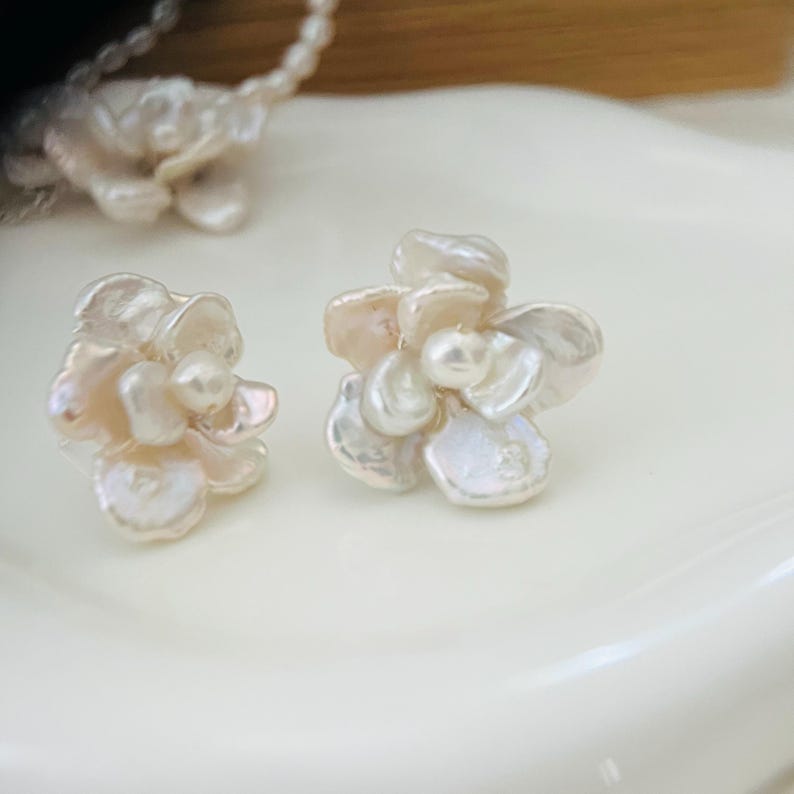 Op de afbeelding: Een paar bloemvormige oorbellen gemaakt van witte en lichtroze parels. De oorbellen liggen op een wit oppervlak, met een bijpassende ketting op de achtergrond. De oorbellen hebben een delicaat, vrouwelijk ontwerp.
