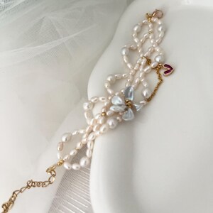 May include: A delicate pearl bracelet with a gold chain and clasp. The bracelet features small, irregular pearls, with a cluster of light blue stones and a small red heart charm. The bracelet is displayed on a white surface.